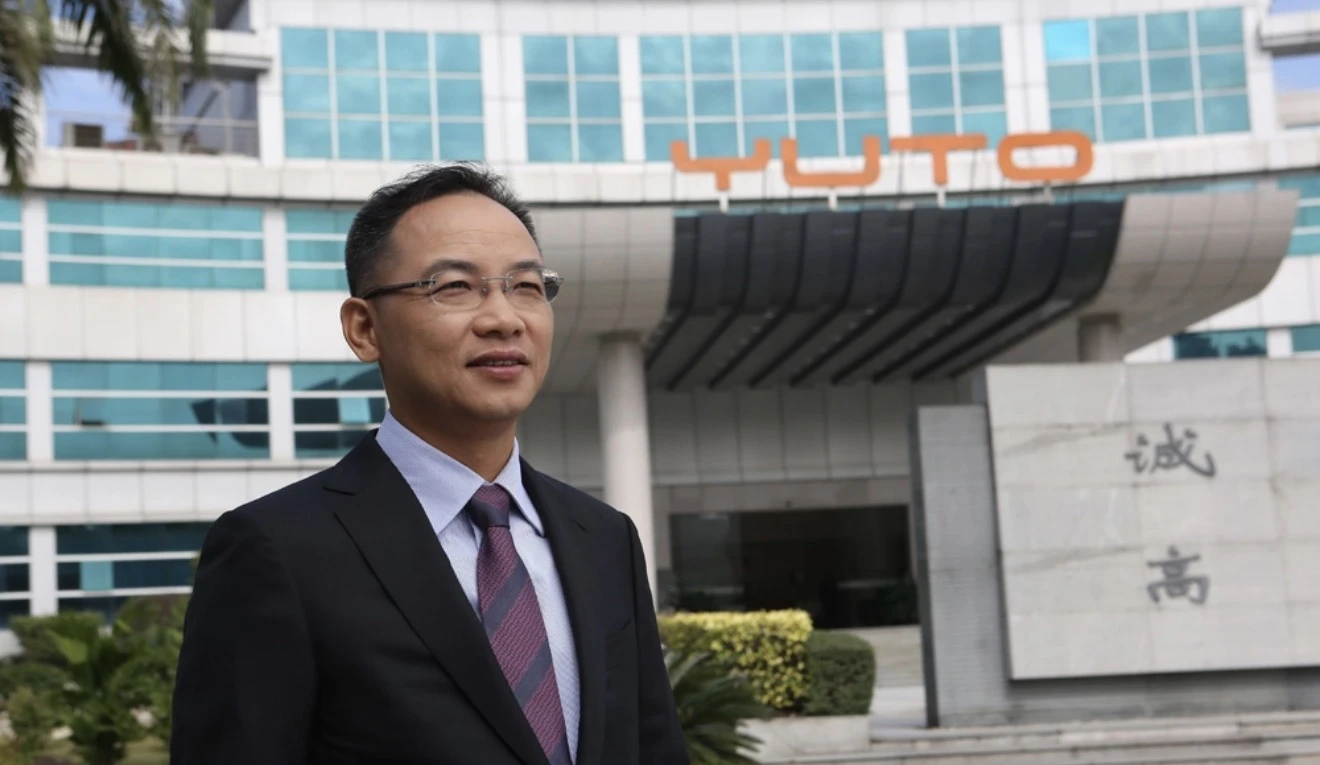 Reference photograph: a person wearing glasses and a dark suit stands before a corporate building bearing the YUTO logo.