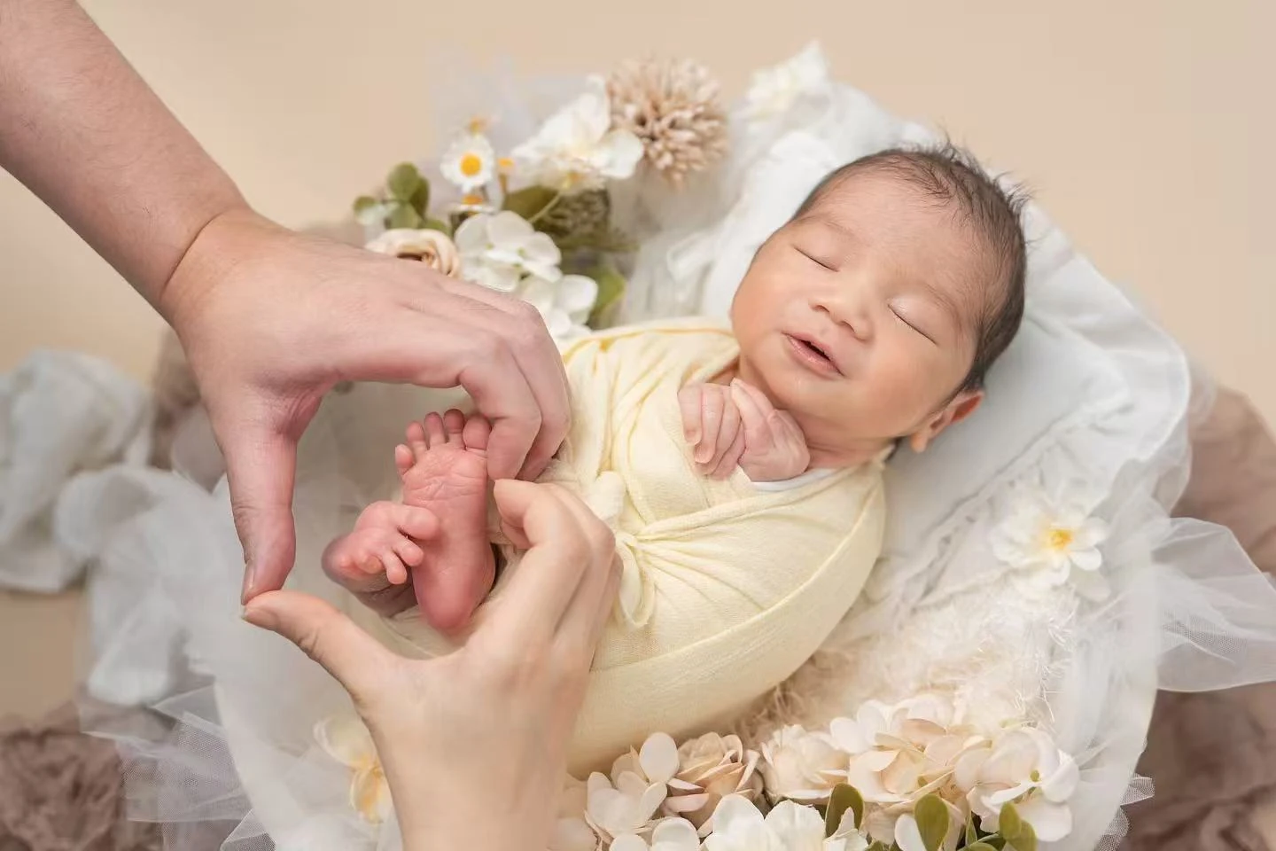 A newborn sleeps among pale fabric and flowers, with hands around tiny feet.