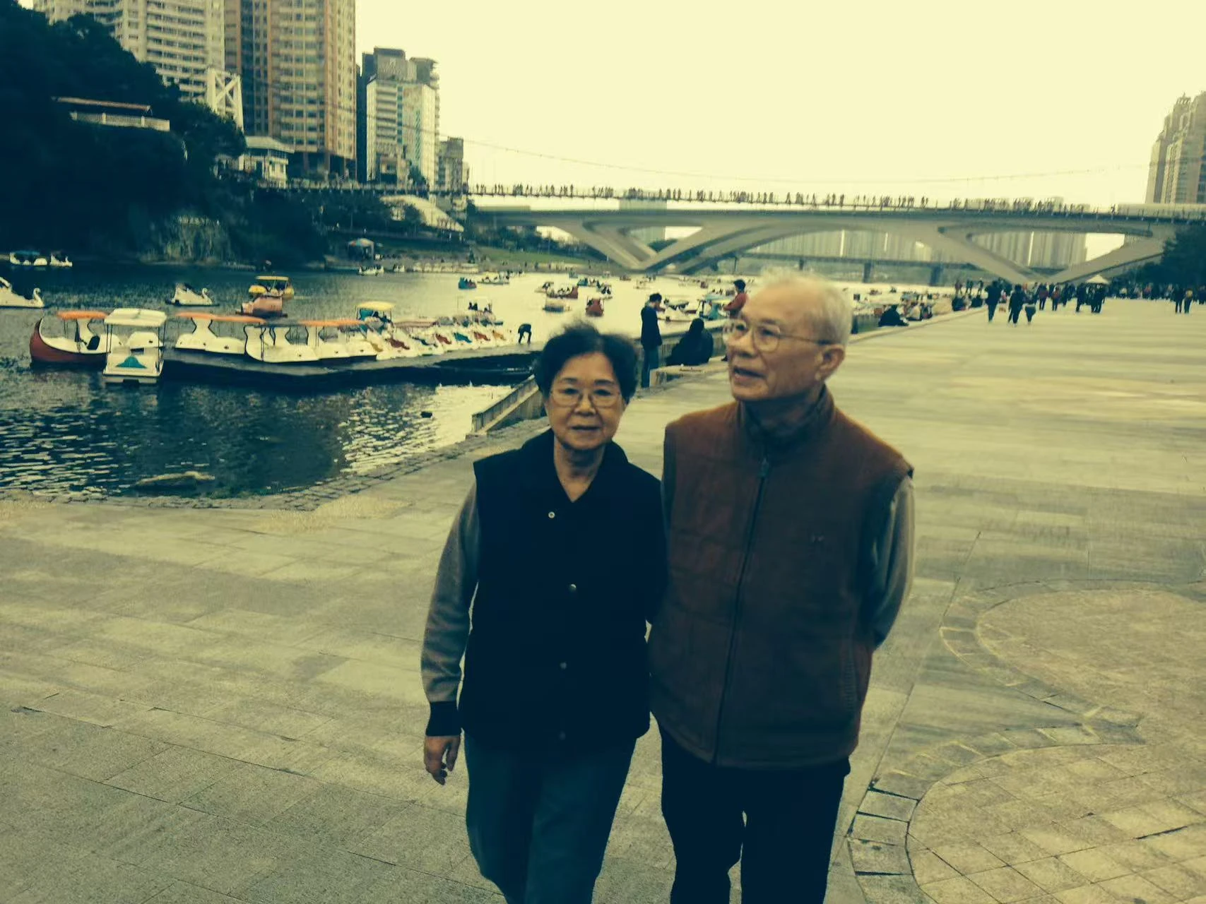 Two people stand side by side on a riverbank, with boats and a bridge behind them.