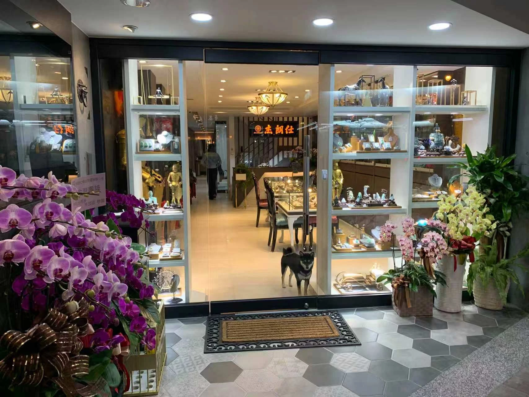 Flowers surround a shop entrance, with glass display cabinets and a dog beside the door.