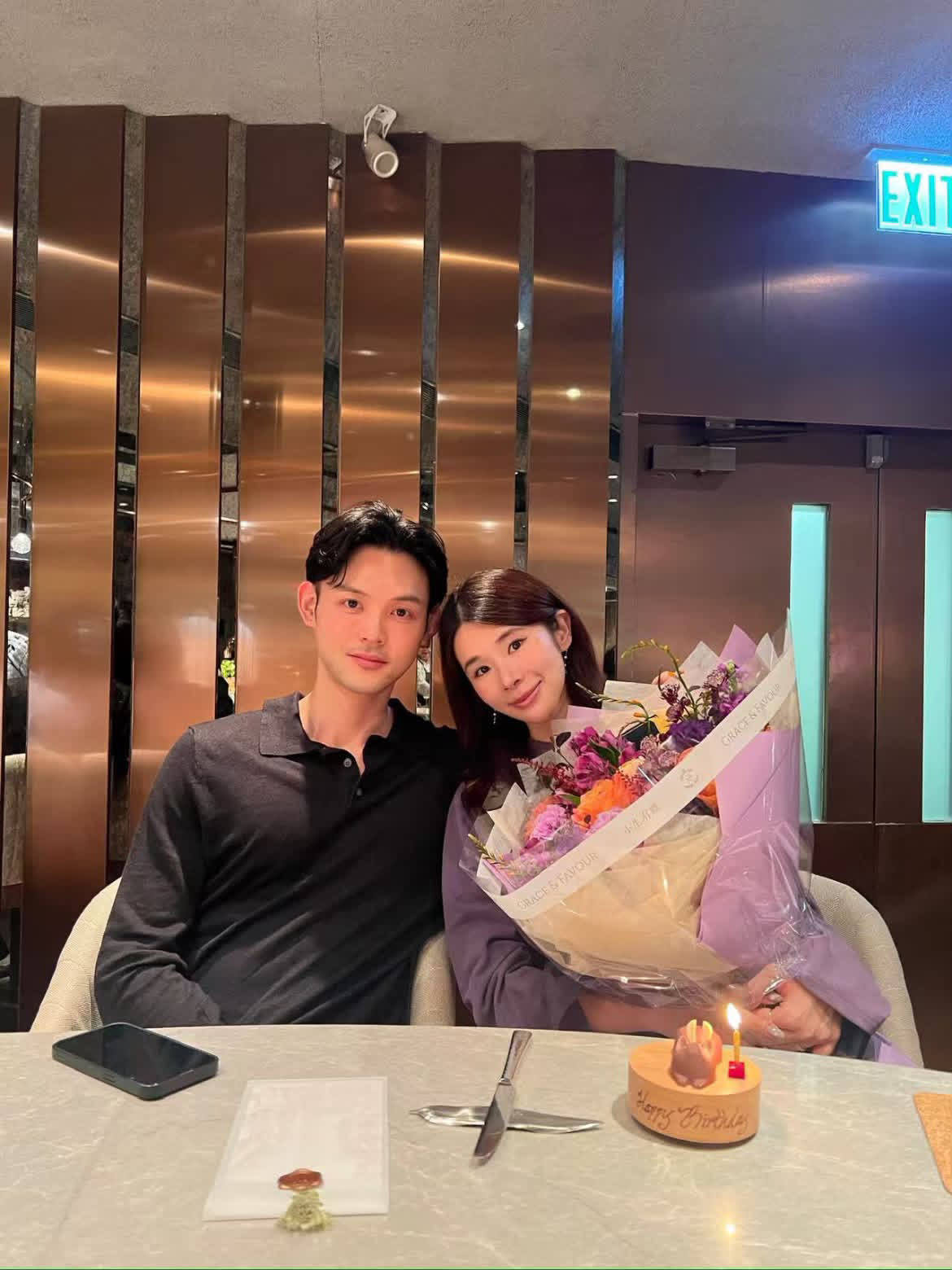 兩人並肩坐在餐桌前，女子抱著花束、靠向身旁的人，桌上有點著蠟燭的生日蛋糕。