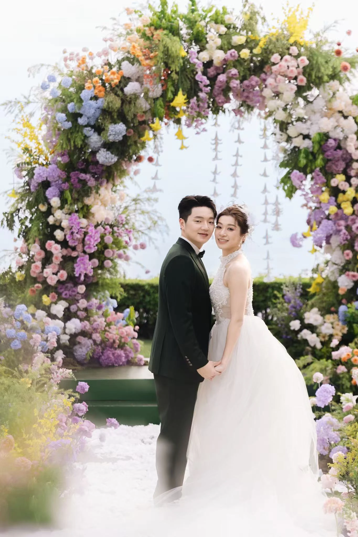 元の結婚写真:黒い礼服と白いドレスの二人が、色鮮やかな花のアーチ前で寄り添い、手をつなぐ。