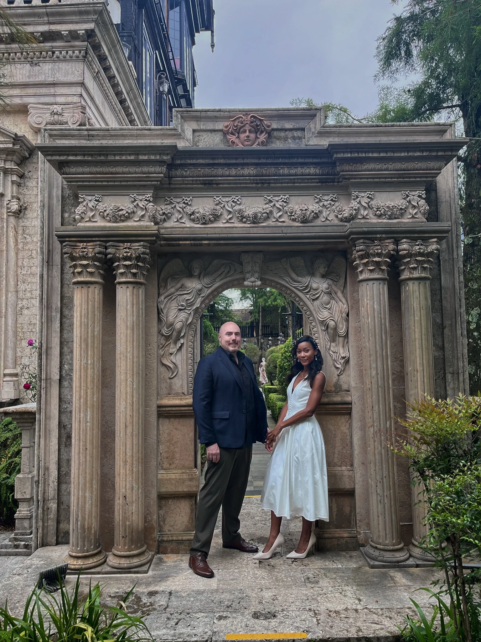 Original proposal photograph: a couple holds hands before a carved stone arch, with garden greenery behind them.